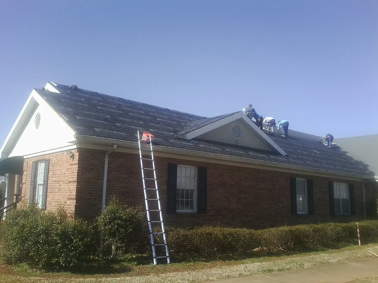 Crew tearing shingles off a brick ranch roof with a ladder set — Emergency Roof Repair work by our crew in Lumberton